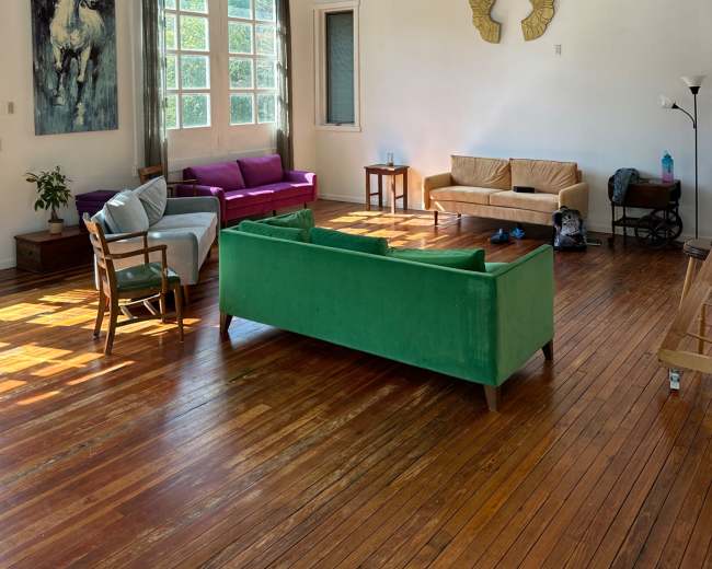 The room features wooden beams on the ceiling, a green sofa, a pink sofa, a brown couch, and large windows allowing natural light to fill the space.