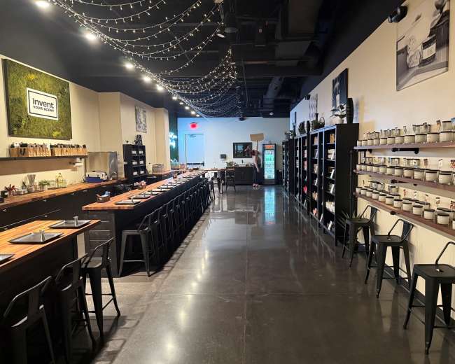 The image shows a modern kitchen space with wooden tables, black stools, and shelves lined with cooking supplies and ingredients under soft string lights.