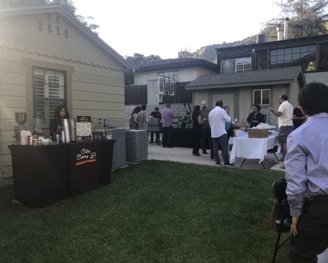 A backyard gathering features a food and beverage table on the left, with guests mingling and dining in an outdoor setting.