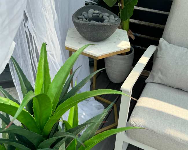 A white curtain drapes beside a gray patio chair and a small table with a decorative bowl, surrounded by large green plants.