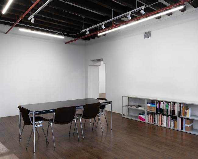 The image shows a minimalistic meeting room with a long table, four chairs, and a shelf filled with books against a white wall.