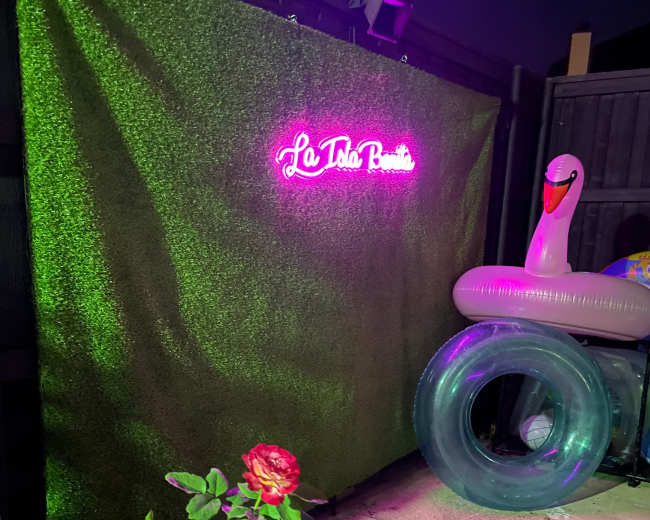 A green backdrop with a neon sign that reads "La Tola Bonita," alongside an inflatable swan and pool rings.