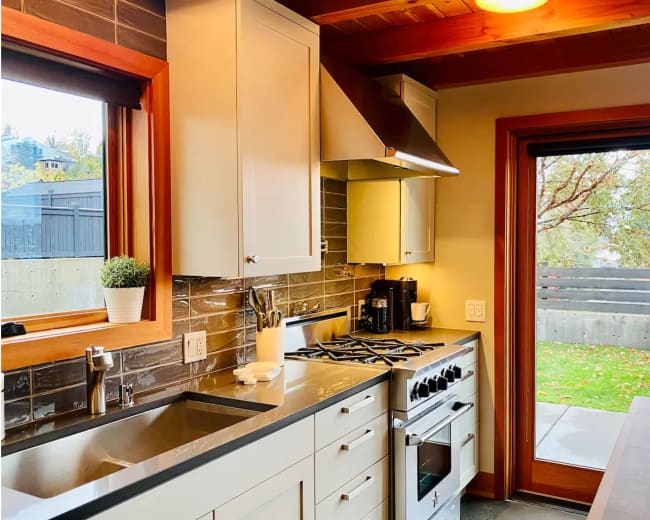 The image shows a modern kitchen with grey cabinets, a stainless steel range hood, and large windows overlooking a patio area.