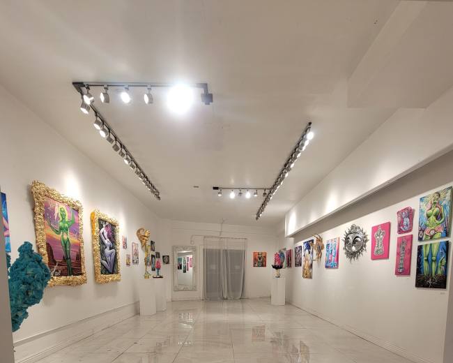 The image depicts a modern art gallery with white walls, polished floors, and various colorful artworks displayed on the walls and on pedestals.