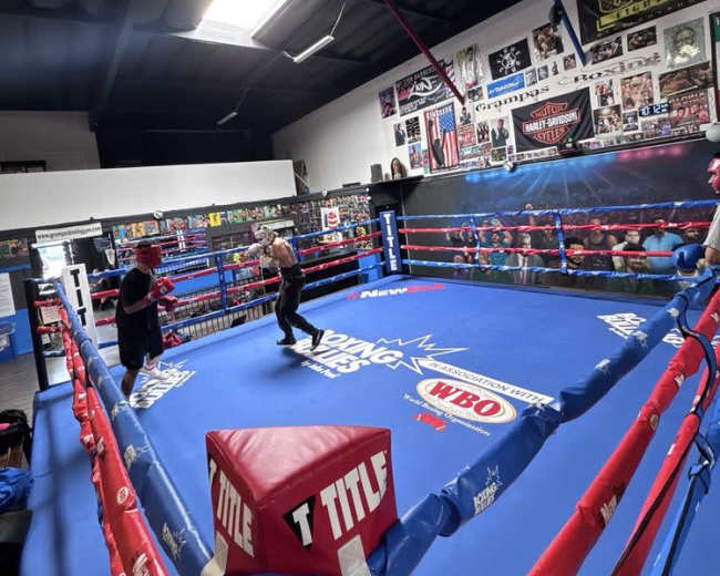 A boxing ring is set up in a gym, surrounded by spectators and decorated with various posters and banners.