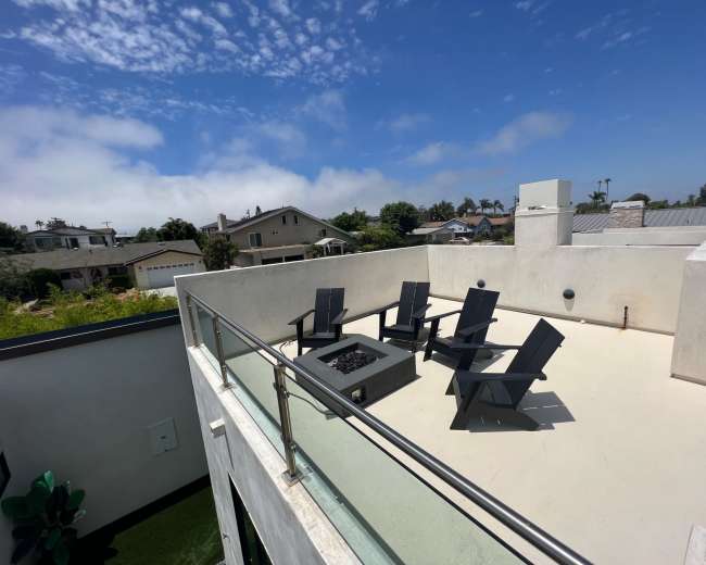 An outdoor rooftop area featuring a fire pit surrounded by five black chairs under a partly cloudy sky.