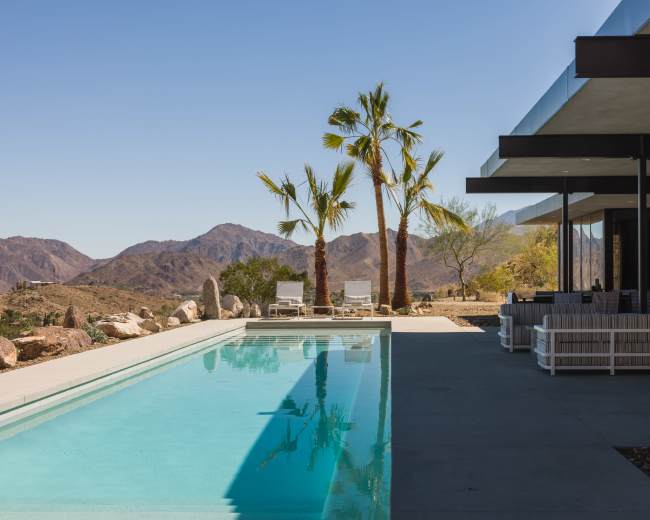 A modern house with a swimming pool is situated against a backdrop of mountains and palm trees.