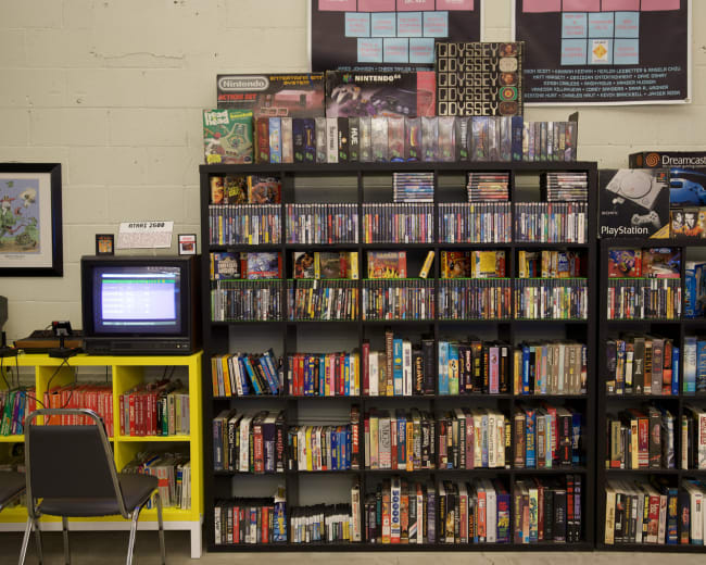A well-organized display of video games and consoles on shelves, with a retro TV and chairs in the foreground.