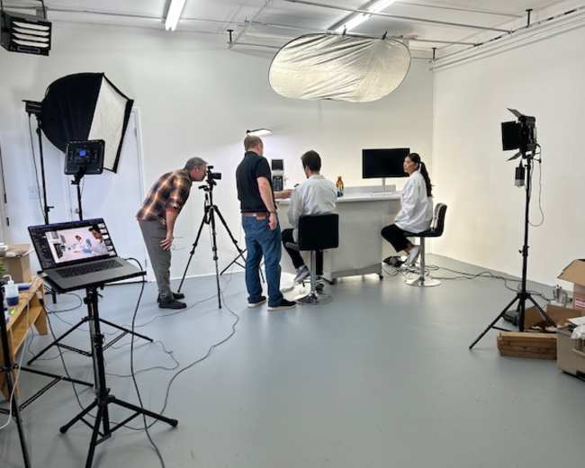 A video production team is set up in a studio, with two individuals seated at a table and cameras positioned to capture the scene.