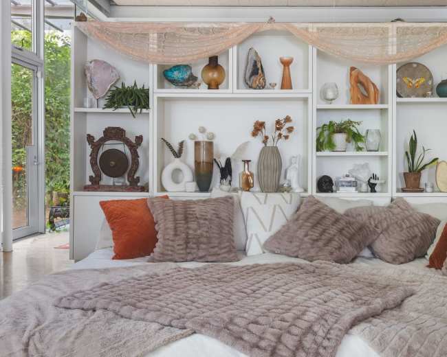 A cozy seating area features a bed with various textured pillows, draped with a soft blanket, in front of a decorative shelf filled with plants and art objects.