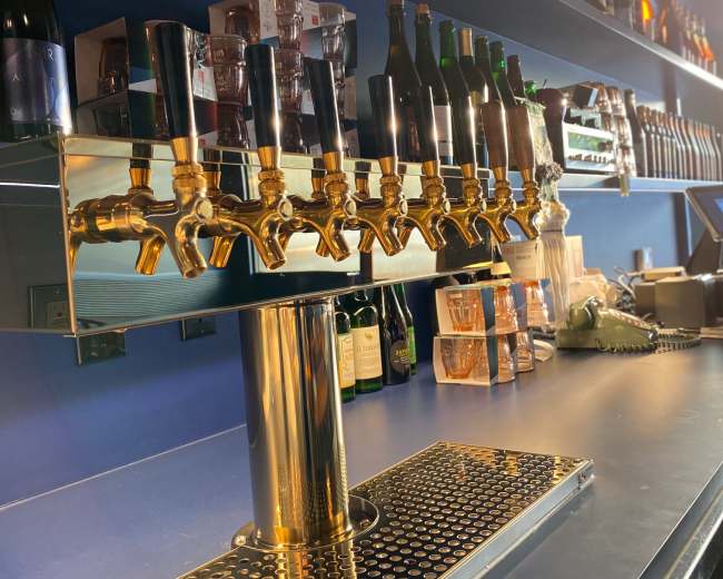 A polished bar with a row of eight shiny brass beer taps and shelves lined with various bottles in the background.