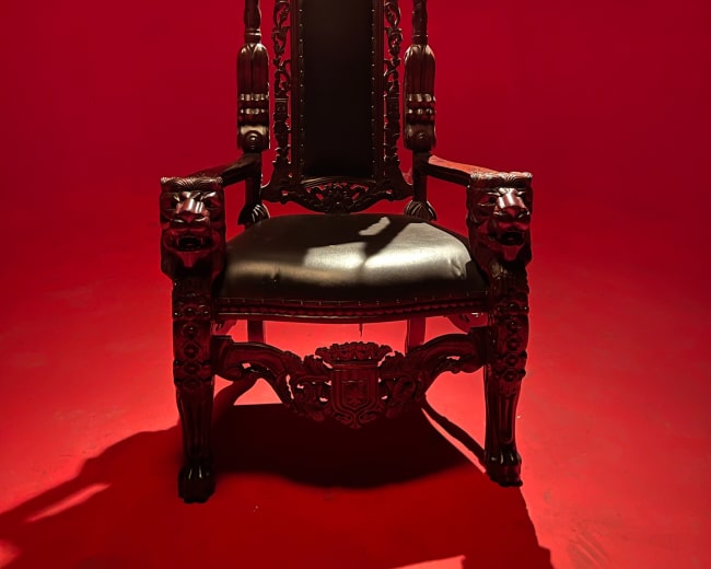 A tall, ornate wooden throne with lion head armrests is positioned in front of a solid red backdrop.