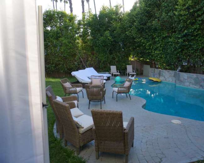 A backyard pool area features a stone patio surrounded by several wicker chairs and lounge seating, with lush greenery in the background.