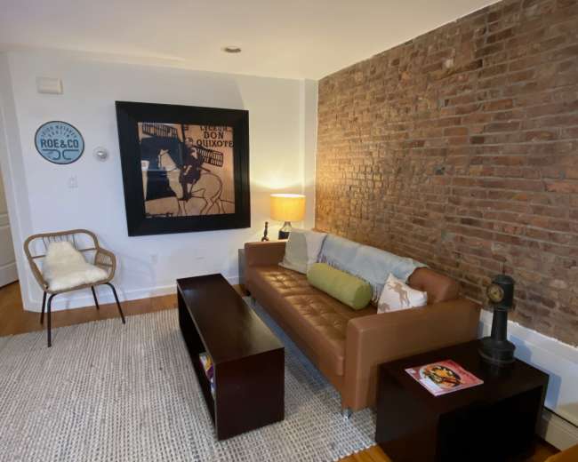 A cozy living room featuring a brown leather sofa, a wooden coffee table, a chair, and a textured rug against a brick wall.