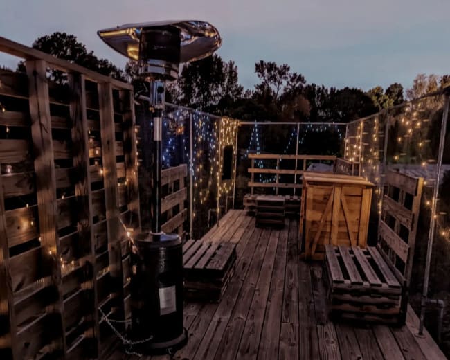 A wooden rooftop deck features string lights, seating areas made from pallets, and a patio heater.