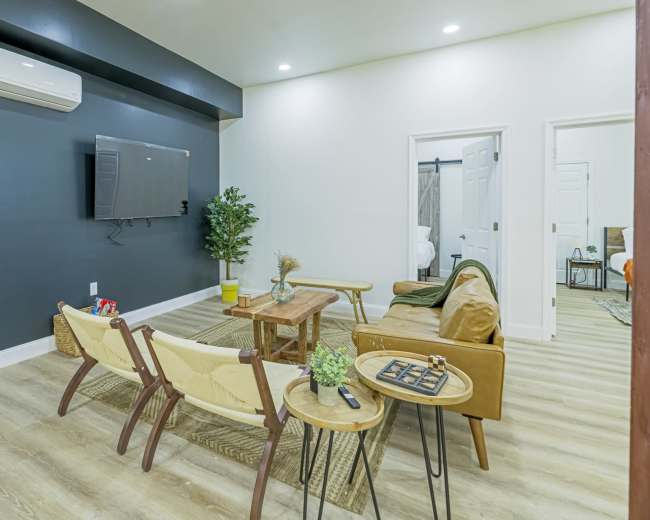 The image displays a modern living room with a television mounted on a black wall, a wooden coffee table, and two armchairs arranged around it.