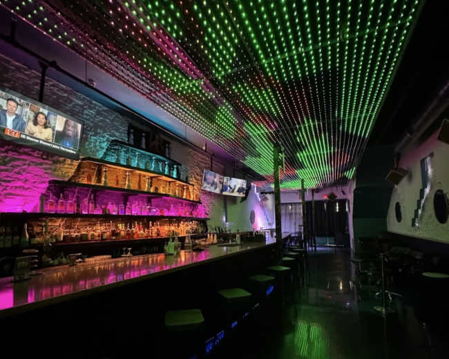 The image shows a modern bar with a colorful LED ceiling, illuminated shelves behind the bar, and multiple screens displaying content.
