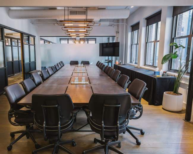 A large conference room features a long wooden table surrounded by ergonomic black chairs, with natural light coming through windows and modern lighting overhead.