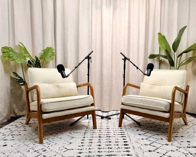 The image shows two mid-century modern chairs facing each other with microphones on stands positioned between them, set against a light curtain and a patterned rug.