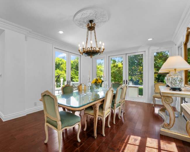 The image shows a well-lit dining room featuring a glass table surrounded by six green upholstered chairs, with large French doors that open to a patio area.