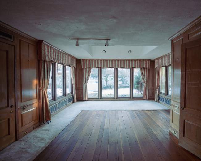 The image shows an empty room with wooden floors, large windows, and wooden paneling along the walls.