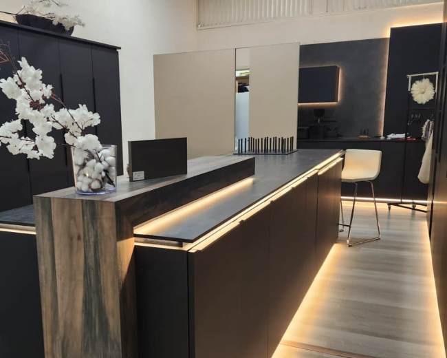 The image shows a modern kitchen design featuring dark cabinetry with under-cabinet lighting and a sleek countertop, complemented by a minimalist decor.