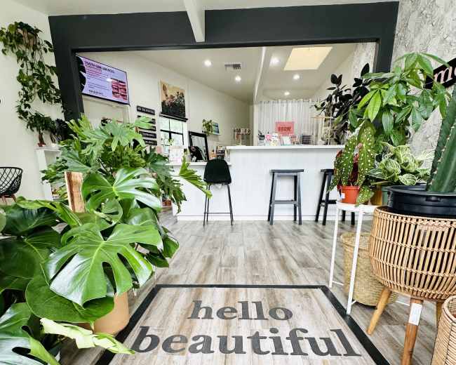 The image shows an indoor space with a welcoming entrance featuring a wooden floor marked with the text "hello beautiful," surrounded by various plants and a bar area in the background.