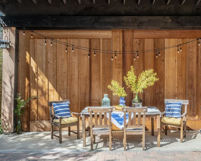 A wooden patio features a table set with a blue and white tablecloth, surrounded by three chairs and decorated with a vase of greenery.
