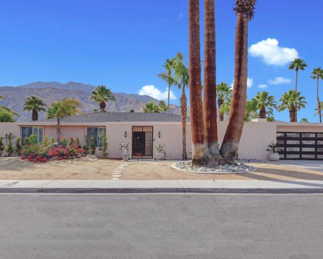 A single-story residential home with palm trees and a desert landscape sits at the end of a sidewalk, against a backdrop of mountains.