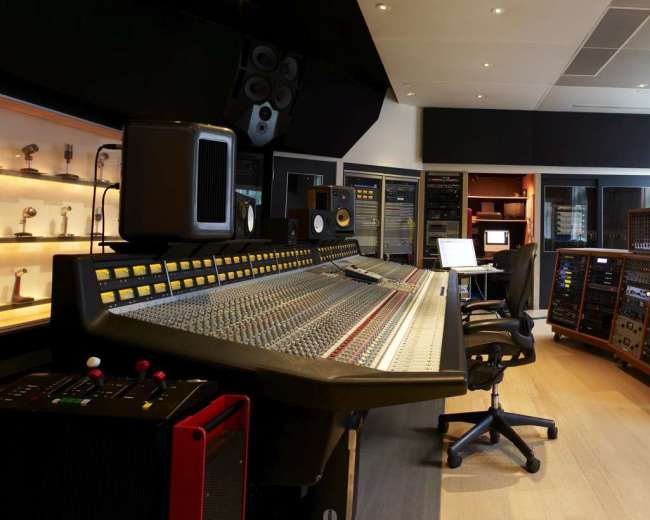 The image shows a recording studio with a large mixing console, several audio equipment racks, and a guitar displayed in the corner.