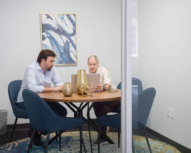 Two people work together at a round table in a well-lit conference room, with a laptop and decorative items in the center.