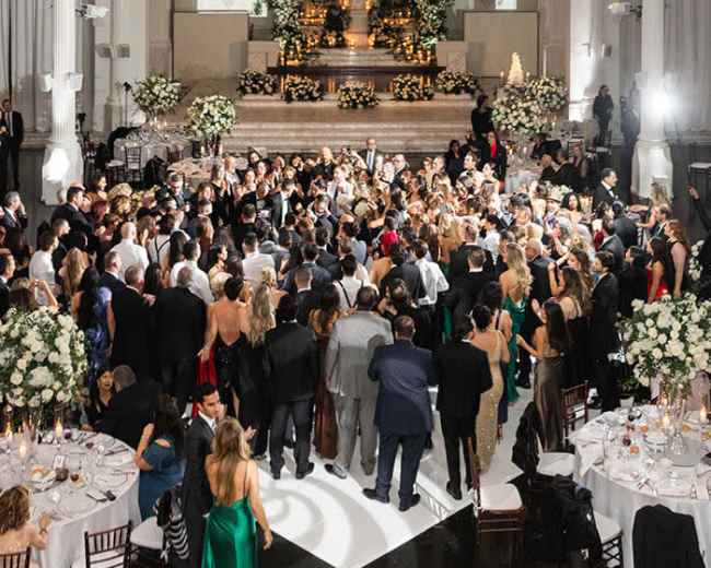 A large crowd gathers in an elegantly decorated venue with a projected silhouette of a couple on the wall.