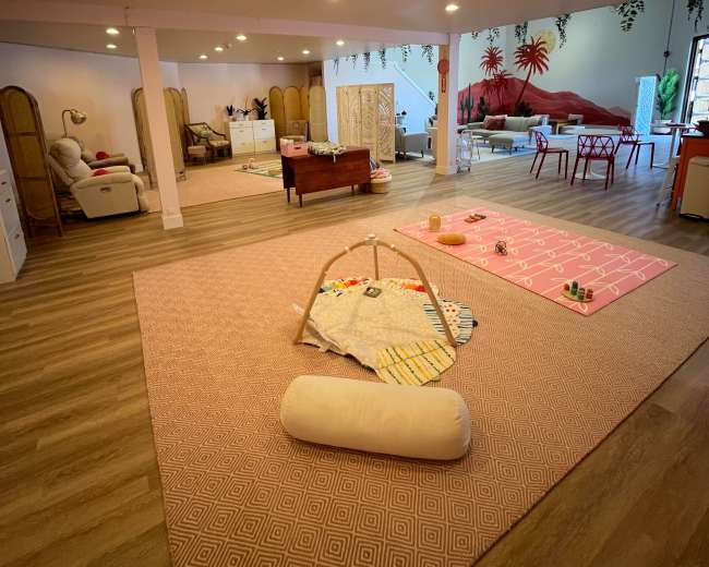 The image shows a spacious, well-lit playroom with various toys, mats, and furniture arranged on wooden flooring.