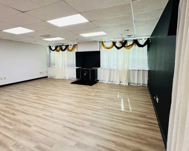 The image shows a spacious, empty room with wooden flooring, a television set on a stand, and curtains with decorative ties at the windows.