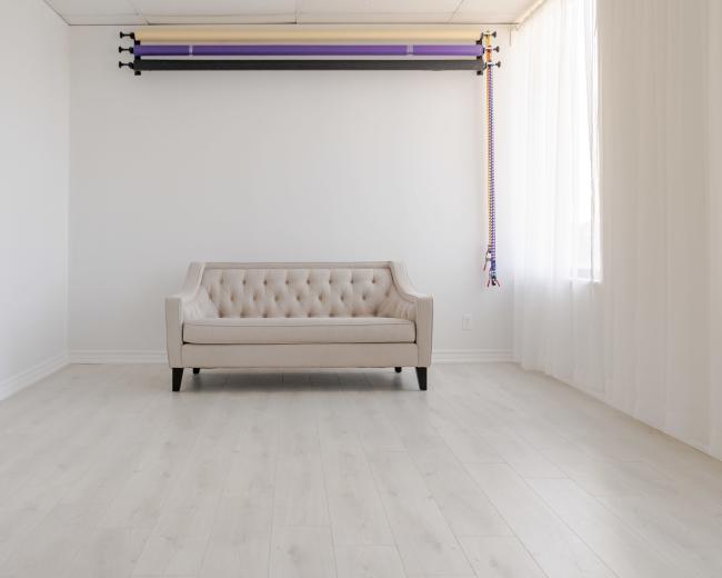A light-colored, tufted sofa is positioned in the center of a spacious, empty room with white walls and a large window.