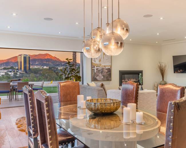 A dining area features a round glass table surrounded by upholstered chairs, with a backdrop of large windows showing a mountain view and pendant lighting above.