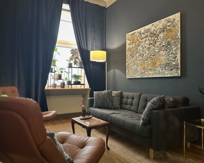 The image shows a cozy living room featuring a dark blue wall, a gray sofa, a wooden table, and large windows with plants.