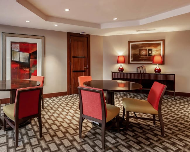 The image shows a modern interior space featuring two round wooden tables surrounded by colorful chairs, with artwork and lamps illuminating the room.
