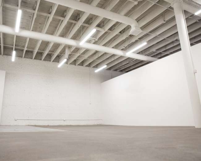 The image shows a sparse, empty room with concrete flooring and white walls, illuminated by overhead lights.