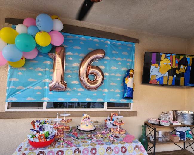 A decorated table with a birthday cake and party snacks is set up under a banner displaying "16," accompanied by balloons and a TV showing animated characters.