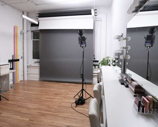 The image shows a well-lit photography studio with a gray backdrop, two light stands, and a vanity area featuring mirrors and makeup products.