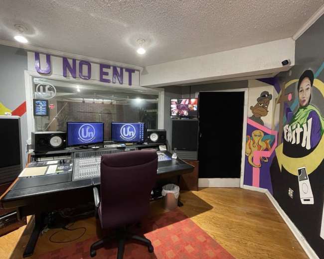 The image shows a music recording studio with a control desk equipped with speakers, a central mixing console, and colorful murals on the walls.