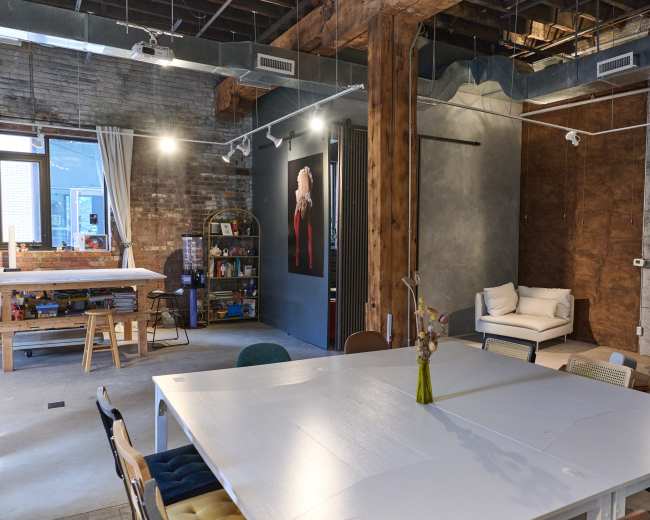 The image shows a modern workspace with a large white table surrounded by multi-colored chairs, a lounging area, and exposed brick and wood elements.