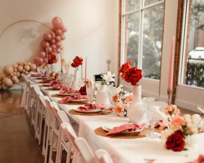 A long dining table is set with decorative plates, napkins, and floral arrangements, flanked by white chairs and a backdrop of pink balloons.