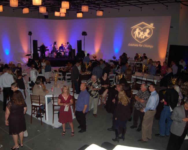 A large group of people socializes at a formal event, featuring a band performing on an elevated stage against a lighted backdrop.