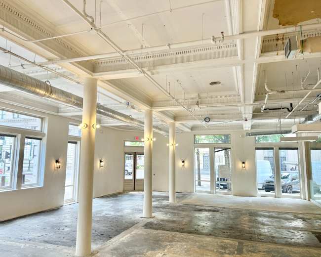 The image shows an empty, well-lit commercial space with large windows, white walls, exposed pipes, and wooden columns.