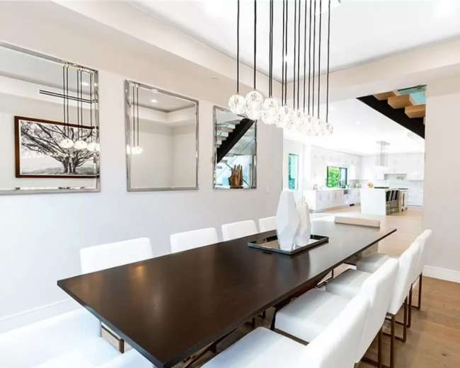 A modern dining room features a long dark wood table surrounded by white chairs, with pendant lighting above and large mirrors on the walls.