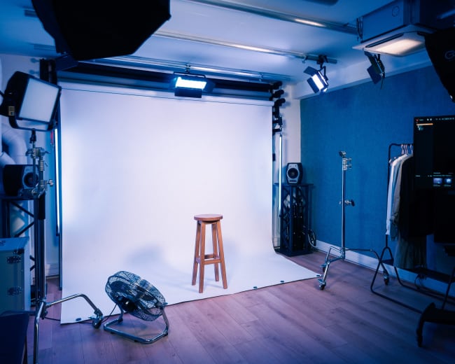 A photography studio with a wooden stool positioned in front of a white backdrop, surrounded by studio lights and various equipment.