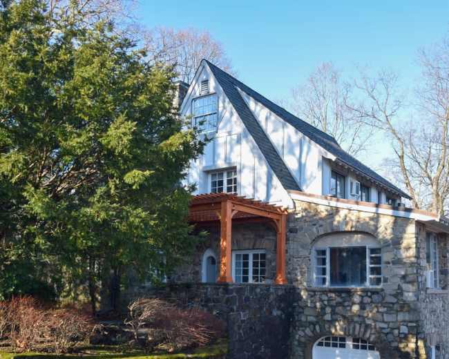 A two-story house with a stone base, wooden porch, and a sloped roof, situated among bare trees.