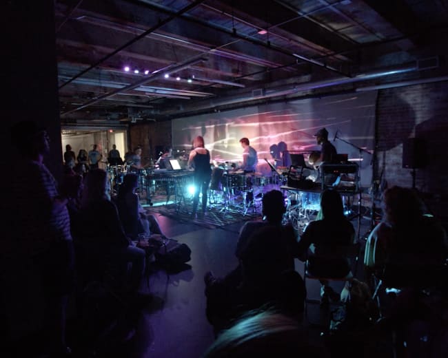 A crowd watches musicians perform onstage in a dimly lit venue with colorful lighting and various instruments.